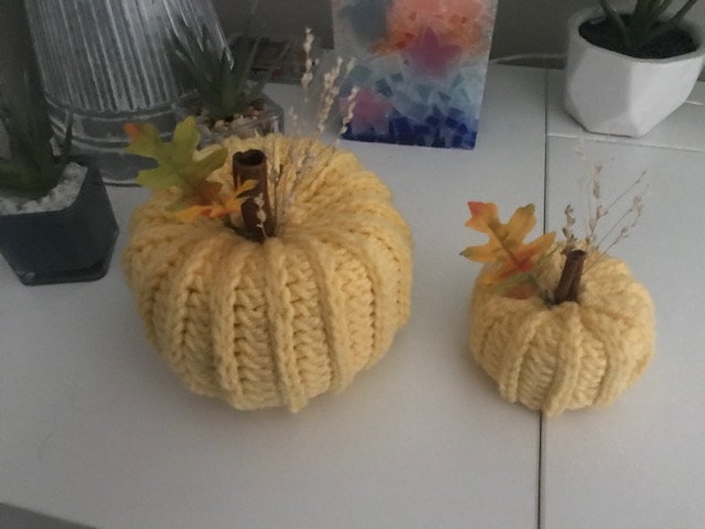 K&ouml;nnte beinhalten: Zwei gestrickte K&uuml;rbisse, einer gro&szlig; und einer klein, in gelber Farbe. Die K&uuml;rbisse haben einen Stiel aus Zimtstange und Herbstdekorationen.