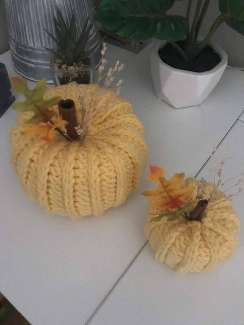 K&ouml;nnte beinhalten: Zwei gestrickte K&uuml;rbisse in gelber Farbe mit Herbstlaub-Akzenten. Der gr&ouml;&szlig;ere K&uuml;rbis hat einen Stiel aus Zimtstange und der kleinere K&uuml;rbis hat einen Stiel aus braunem Zweig.