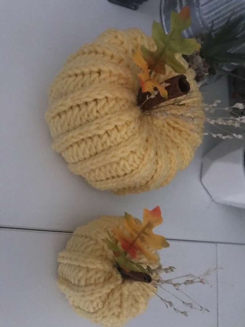 K&ouml;nnte beinhalten: Zwei gestrickte K&uuml;rbisse mit Herbstdekoration. Der gr&ouml;&szlig;ere K&uuml;rbis ist gelb und hat einen Zimtstange und Herbstbl&auml;tter. Der kleinere K&uuml;rbis ist ebenfalls gelb und hat einen Zimtstange und Herbstbl&auml;tter.