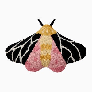 May include: A moth-shaped cushion with a detailed design. The wings are black with white accents, and the body features pink, yellow, and cream colours. The cushion has a textured, tufted appearance and is set against a white background.