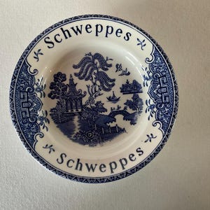 May include: A white ceramic plate with a blue and white willow pattern. The plate has the word "Schweppes" written around the rim.