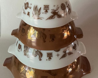 May include: A stack of four nesting bowls with handles, alternating between white and brown glass. Each bowl features a repeating metallic gold pattern of roosters, corn, coffee grinders, and small star shapes around the exterior surface.