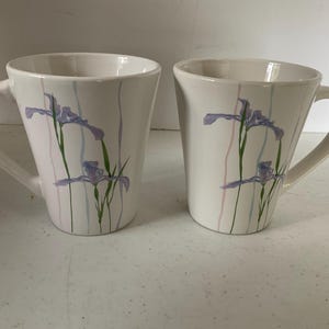 May include: Two white ceramic mugs with a floral design. The mugs feature a light purple iris flower pattern on a white background with thin pastel stripes.