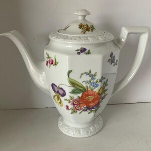 May include: A white porcelain teapot with a floral design. The teapot has a octagonal body and a lid with a knob. The flowers are in shades of pink, purple, yellow, and orange.