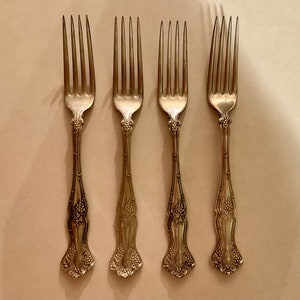 May include: Four silver forks with ornate designs on the handles. The forks are arranged in a row on a white surface.