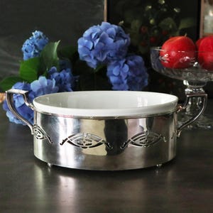 May include: A silver serving dish with a white porcelain insert. The dish has a decorative cut-out design around the rim and two handles.