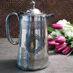 May include: An antique silver coffee pot with a lid and handle. The pot has decorative detailing, including an oval emblem and patterned bands. A bouquet of pink tulips and white flowers is in the background.