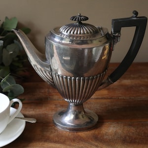 May include: A silver teapot with a black handle and a fluted design. The teapot has a round body and a pedestal base.