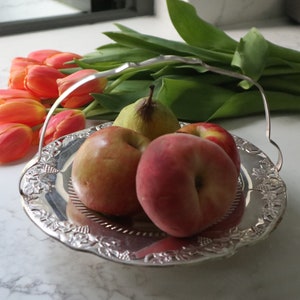 Op de afbeelding: Een zilveren serveerschaal met een sierlijk ontwerp heeft een selectie fruit, waaronder rode appels, perziken en een groene peer. De schaal heeft een handvat en is omringd door een boeket oranje tulpen.