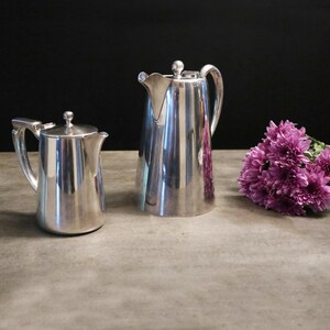 May include: Two silver pitchers with curved handles and lids. The larger pitcher has a spout with a decorative edge. A bouquet of purple flowers is to the right of the pitchers. The background is black.