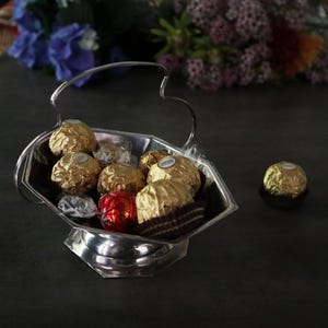 May include: A silver serving dish with a handle, filled with assorted chocolates wrapped in gold foil. The chocolates are arranged in a decorative pattern.