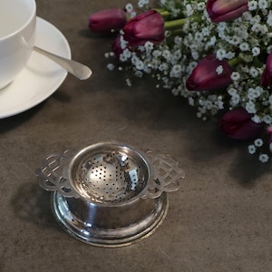 May include: A silver tea strainer with a decorative handle rests on a matching base. A white teacup and saucer with a spoon are in the background. A bouquet of purple tulips and white flowers is also visible, creating a classic tea setting.