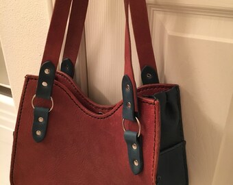 May include: A small, brown leather handbag with a blue leather pocket on the side. The bag has two straps and a top zipper closure.