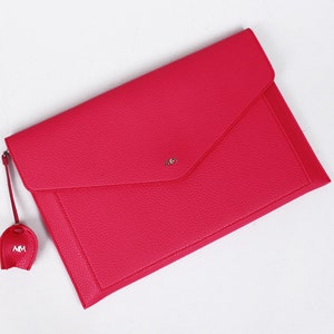 May include: A bright pink leather envelope clutch with a small, matching pink tassel. The tassel has the letters "MM" in silver.