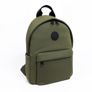 May include: Olive green backpack with a rounded shape, a front pocket, and a top handle. The backpack has black zippers and a circular black patch with white text. The bag is made of a smooth, durable material.