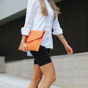 May include: A woman wearing a white button-down shirt and black biker shorts is holding an orange leather clutch.
