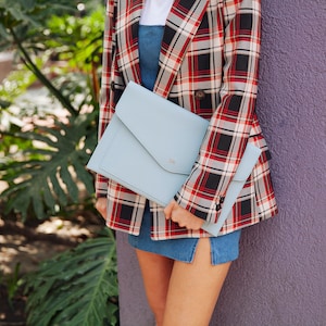 May include: A woman is holding a light blue leather envelope clutch with a gold monogram. She is wearing a red and black plaid blazer and a denim skirt.