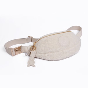 May include: A white faux fur fanny pack with a gold zipper and a gold buckle. The bag has a small, round, white leather patch with a circle logo on the front. A gold chain with a white leather bunny charm is attached to the bag.
