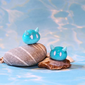 Puede incluir: Dos pequeñas figuritas de cerámica azul con ojos blancos y cuernos, sentadas sobre rocas grises y marrones. Las figuritas tienen forma de personaje de dibujos animados con un cuerpo redondeado y una cabeza pequeña y puntiaguda.