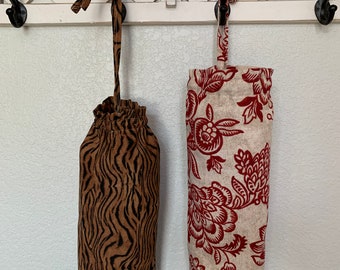 May include: Two fabric bags for holding plastic grocery bags. One bag is brown with black tiger stripes and the other is beige with red floral designs. Both bags are hanging from hooks on a white wall.