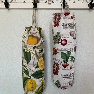 May include: Two fabric bags with a drawstring closure hang on hooks. The bag on the left has a lemon and green leaf pattern. The bag on the right has a red tomato, beet, radish, and garlic pattern. Both bags have text that reads "Garlic Delicious Homegrown & Handpicked" and "Beet 12 Varieties".