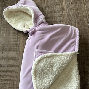 May include: A light purple baby carrier cover with a hood and a soft, white, fluffy interior. The cover has a button closure and a blanket-like section with a white, textured lining. The exterior is a smooth, solid color.