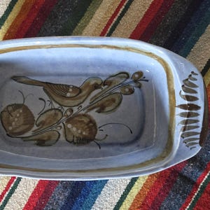 May include: A rectangular ceramic dish with a light blue interior and a brown bird and branch design. The dish has a brown border and handles. The dish is resting on a striped red, yellow, and green textile.