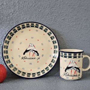 May include: A ceramic plate and mug set with a stork design. The plate and mug are cream-colored with a dark blue border and feature a stork illustration. The plate is approximately 8 inches in diameter, and the mug has a handle.