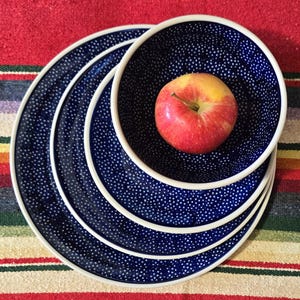 May include: A stack of blue ceramic plates and a bowl with a white rim and a white dotted pattern. A red and yellow apple sits inside the bowl. The plates and bowl are on a colorful striped surface.
