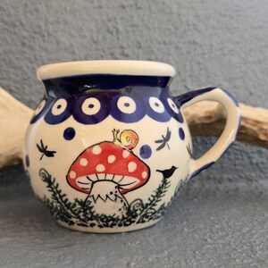 May include: A round ceramic mug with a cream-colored base featuring a hand-painted red and white spotted mushroom topped with a small snail. The design includes black silhouettes of dragonflies and a bird, with a dark blue rim and handle.