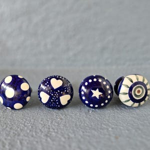 May include: Four decorative ceramic cabinet knobs in a row. The knobs are round and painted in navy blue and white. Designs include polka dots, hearts, a star, and a floral pattern. These knobs are perfect for adding a touch of style to any cabinet or drawer.