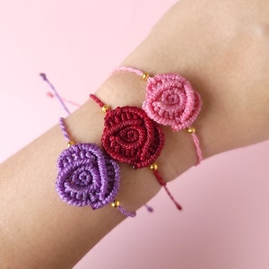 May include: Three friendship bracelets with a rose design. The bracelets are made with purple, red, and pink string and have gold beads.
