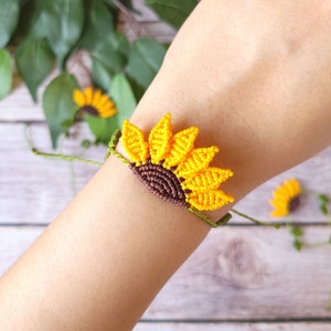 May include: A handmade macrame bracelet featuring a yellow sunflower design. The sunflower has yellow petals and a brown center. The bracelet is attached to a green cord. A small sunflower is visible in the background.