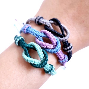 May include: Four friendship bracelets in different colors, including teal, purple, blue, and black, are stacked on a person's wrist. The bracelets are made of knotted string and have a simple, elegant design.