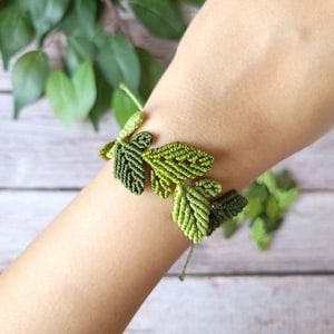 May include: A green macrame bracelet with a leaf design. The bracelet is made with a variety of shades of green cord.