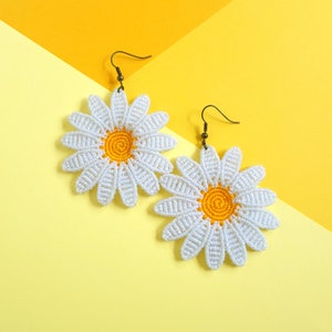 May include: A pair of white crocheted daisy earrings with yellow centers. The earrings are on a yellow and light yellow background.