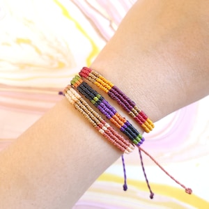 May include: A set of five colorful friendship bracelets with a woven pattern. The bracelets are made of thin cords in shades of purple, orange, red, gold, and green.
