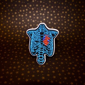 Puede incluir: Una pegatina azul y blanca de un esqueleto con un corazón brillante. La pegatina está sobre un fondo negro con estrellas doradas.