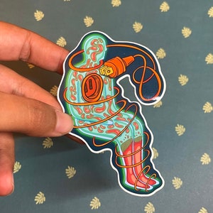 May include: A sticker of a green and orange figure with a cord wrapped around it. The figure is sitting with its legs crossed and has a plug in its chest.