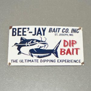 Op de afbeelding: Een rechthoekig wit bord met de tekst "BEE'-JAY BAIT CO. INC" en "DIP BAIT" in het rood. Het bord toont een blauwe meerval illustratie en de zin "THE ULTIMATE DIPPING EXPERIENCE".