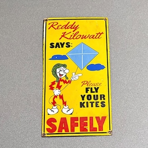 May include: Vintage yellow metal sign featuring Reddy Kilowatt, a cartoon character, holding a blue kite. The sign reads "Reddy Kilowatt SAYS: Please FLY YOUR KITES SAFELY" in bold, black letters. The sign has a distressed look.