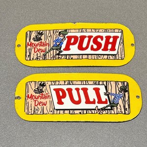 May include: Two yellow metal signs with a woodgrain background. The top sign says "PUSH" in red letters and features a cartoon character pushing a door. The bottom sign says "PULL" in red letters and features a cartoon character pulling a door. Both signs feature the Mountain Dew logo.