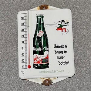 May include: Vintage metal thermometer featuring a Mountain Dew bottle illustration. The thermometer shows Celsius readings and the text "there's a bang in ever' bottle!" and "Dee-lishus Soft Drink!"
