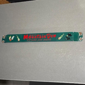 May include: A long, rectangular green metal sign featuring the red text Mountain Dew and the phrase It'll tickle yore innards. The sign includes illustrations of a cartoon character, a glass bottle, and a small black bear graphic on the ends.