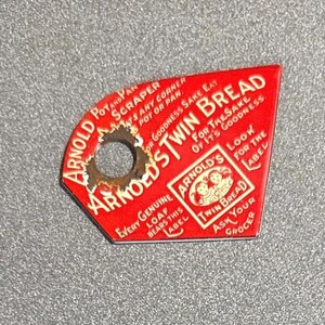 May include: A vintage red Arnold's Twin Bread scraper with a hole for hanging. The scraper features white text that reads "Arnold's Twin Bread" and other text. The scraper is on a gray surface.