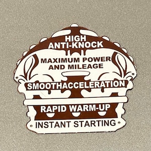 Puede incluir: Un letrero de metal blanco con letras marrones. El letrero tiene forma de corona y tiene el texto "HIGH ANTI-KNOCK MAXIMUM POWER AND MILEAGE SMOOTHACCELERATION RAPID WARM-UP INSTANT STARTING."
