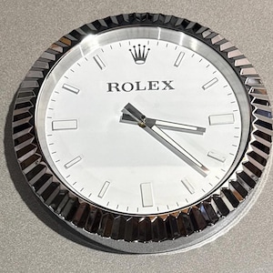 Puede incluir: Un reloj de pared redondo con esfera blanca y manecillas y bisel plateados. La palabra "ROLEX" está impresa en negro en la parte superior central, con el logotipo de la corona Rolex encima. El reloj tiene un bisel estriado.