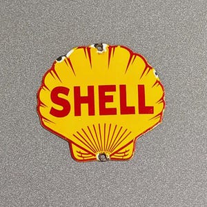 Puede incluir: Un letrero vintage de la compañía petrolera Shell con forma de concha de vieira. El letrero es amarillo con un borde rojo y la palabra "SHELL" en letras rojas en negrita. Tiene dos agujeros para el montaje.