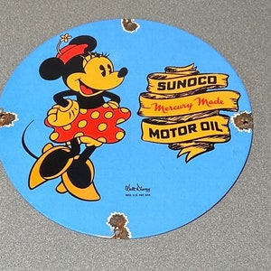 Puede incluir: Un letrero redondo de metal azul y amarillo con una imagen de dibujos animados de Minnie Mouse. El letrero anuncia Sunoco Mercury Made Motor Oil. El texto "Walt Disney" y "REG. U.S. PAT. OFF." está impreso en la esquina inferior derecha.