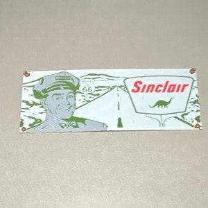 May include: A vintage Sinclair petrol station sign with a green dinosaur logo and the text "Sinclair". The sign also features a man in a uniform and a road with the number "66" on a sign.